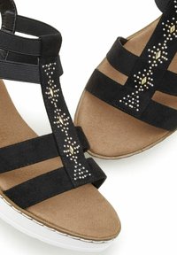 Schwarze Wildledersandalen mit flachem Absatz, ausgestattet mit einem Design aus drei Riemen und verziert mit metallischen Nieten. Gepolstertes, braunes Innenfutter, weiße Sohle.