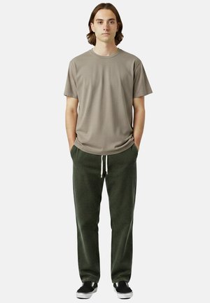 Giovane uomo in piedi con le mani in tasca che indossa una t-shirt color taupe, pantaloni verdi scuri con coulisse e sneakers nere slip-on su sfondo bianco.