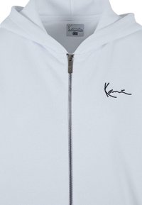 White zip-up hoodie made of soft fabric, featuring a drawstring hood, a silver zipper, and a black embroidered logo on the left chest.