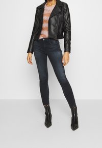 Femme portant une veste en cuir noire, un pull rayé rose et orange, un jean skinny foncé et des bottines noires pointues, debout contre un mur blanc.