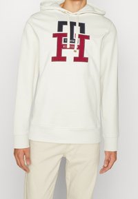 Sudadera con capucha blanca y bolsillo frontal. Presenta gráficos y texto en rojo y azul marino; tejido suave con capucha ajustable. Diseño simple.