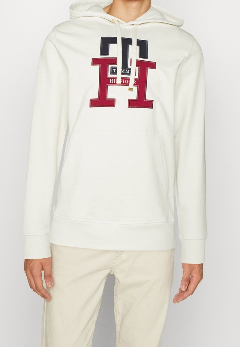 Sudadera con capucha blanca y bolsillo frontal. Presenta gráficos y texto en rojo y azul marino; tejido suave con capucha ajustable. Diseño simple.