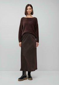 Femme portant un pull bouclé marron foncé, une jupe longue assortie et des bottes noires chunky, se tenant contre un fond gris clair uni.