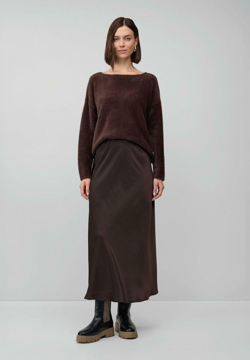 Femme portant un pull bouclé marron foncé, une jupe longue assortie et des bottes noires chunky, se tenant contre un fond gris clair uni.