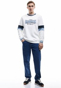 Sudadera blanca con detalles en azul y texto "BROOKLYN", combinada con jeans azules y zapatos negros con cordones blancos y suela de goma.