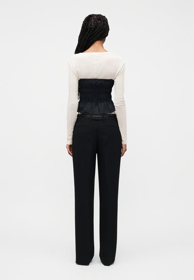 Άτομο με μακριά πλεξούδα που φοράει λευκό μακρυμάνικο πάνω μέρος, μαύρο κορσέ με ρυτιδώσεις και μαύρο παντελόνι με φαρδιά μπατζάκια, στεκόμενο με την πλάτη.