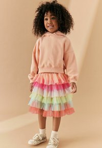 Hoodie pêche, jupe en tulle superposée dans des teintes de rose, vert et bleu, chaussettes blanches et baskets avec accents argentés, sur un fond beige.