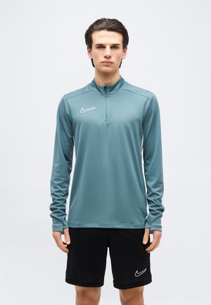Ung mand stående vendt fremad iført en teal långærmet pullover og sorte shorts, begge med hvide Nike-logoer, mod en hvid baggrund.
