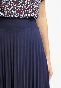 Marinblå plisserad kjol med elastisk midja och slät textur, kombinerad med en mönstrad blus med kort ärm som har bladmönster.