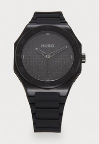 Czarny zegarek z gumowym paskiem i teksturowaną szaro-skalą tarczą z nazwą marki "HUGO". Okrągła koperta, srebrne wskazówki i minimalistyczny design.