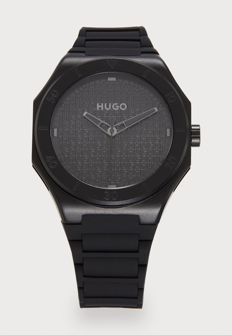 Czarny zegarek z gumowym paskiem i teksturowaną szaro-skalą tarczą z nazwą marki "HUGO". Okrągła koperta, srebrne wskazówki i minimalistyczny design.