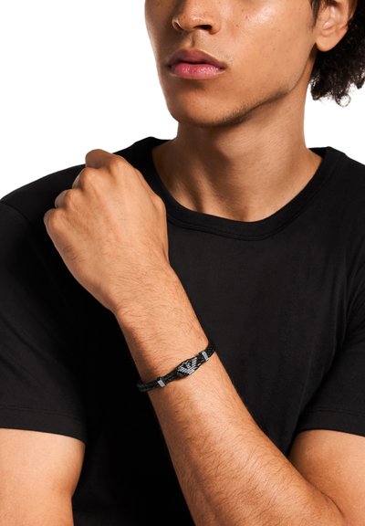 Bracelet tressé noir avec un accent métallique central, porté au poignet. La personne porte un t-shirt noir uni et un fond neutre.