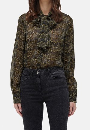 Vrouw die een zwarte doorzichtige blouse draagt met een goudkleurig vuurwerkpatroon en een grote strik bij de hals, gecombineerd met hooggetailleerde zwarte gewassen spijkerbroek.