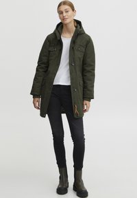 Parka vert olive avec capuche, fermeture à boutons et à zip, et deux poches avant. Portée sur une chemise blanche et un jean noir.