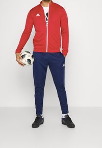 Veste de sport zippée rouge, t-shirt blanc avec un logo, pantalon de survêtement bleu marine, chaussures de sport noires, tenant un ballon de football à motif.