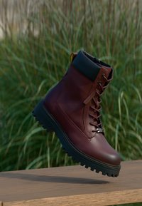 Bota de couro vermelho escuro com atacadores, sola espessa preta, colarinho acolchoado e alça, apresentada no ar acima de uma superfície de madeira com fundo de relva verde.