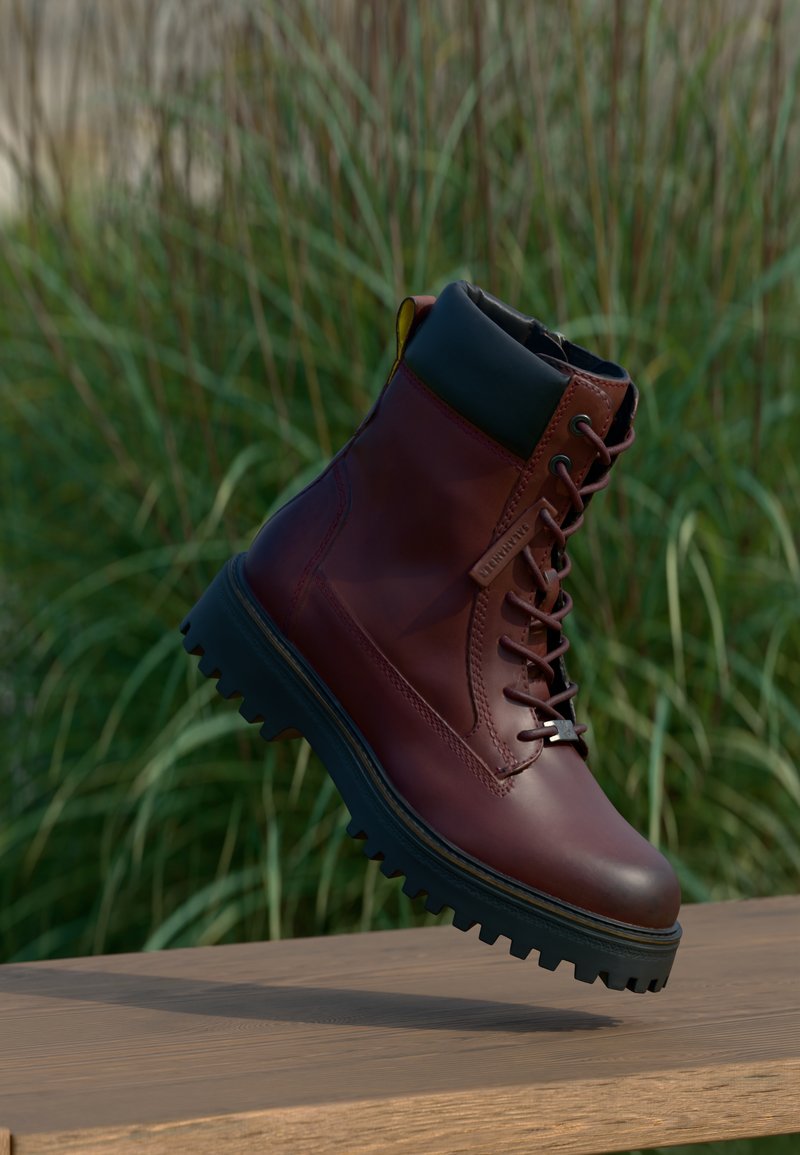 Bota de couro vermelho escuro com atacadores, sola espessa preta, colarinho acolchoado e alça, apresentada no ar acima de uma superfície de madeira com fundo de relva verde.