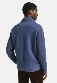 Mann, der sich abwendet, trägt einen blauen Pullover mit hohem Kragen und dunkelbrownen Hosen vor einem schlichten Hintergrund.