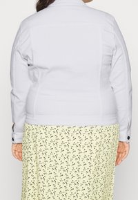 Ljusgrå denimjacka med långa ärmar, knäppta manschetter och rak fåll, bärs över en mönstrad gul kjol med små blommönster.