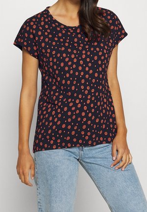Femme portant un t-shirt bleu marine foncé avec des motifs circulaires rouges et un jean taille haute bleu clair, debout devant un fond uni.
