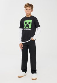 Czarny t-shirt z krótkim rękawem Minecraft z zielonym pikselowym wzorem postaci, noszony na białej koszuli z długim rękawem. Połączony z czarnymi spodniami.