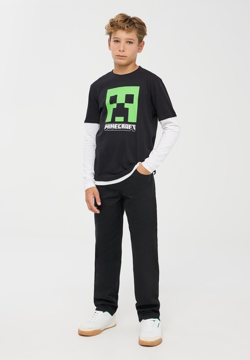 Czarny t-shirt z krótkim rękawem Minecraft z zielonym pikselowym wzorem postaci, noszony na białej koszuli z długim rękawem. Połączony z czarnymi spodniami.