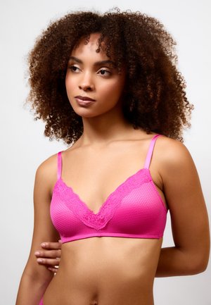 Jeune femme aux cheveux bouclés portant un soutien-gorge rose vif orné de dentelle, regardant légèrement sur le côté sur un fond blanc uni.