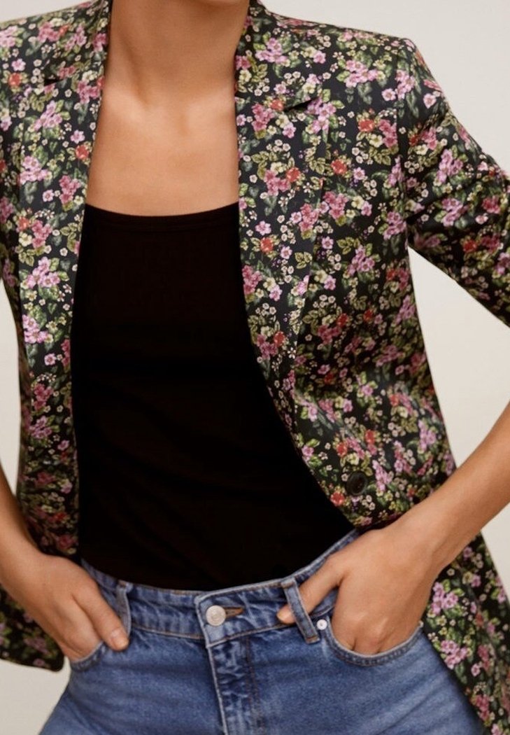 Personne portant un haut noir, un jean bleu et un blazer foncé à imprimé floral, les mains dans les poches du jean.