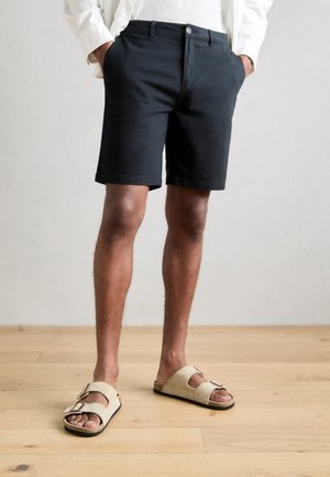 Pantalones cortos negros con una textura suave, con cierre de botón y bolsillos laterales. Se combinan con sandalias beige que presentan un detalle de hebilla.