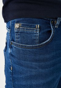 Donkerblauwe spijkerbroek met een klassieke pasvorm, contrasterende gouden stiksels, een zilveren logoknop en vijf zakken. Getextureerde, middelzware stof.