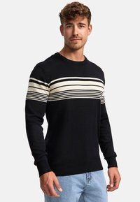 Schwarzer Strickpullover mit horizontalen cremefarbenen Streifen über der Brust, Rundhalsausschnitt sowie gerippten Bündchen und Saum, getragen über hellblauen Jeans.