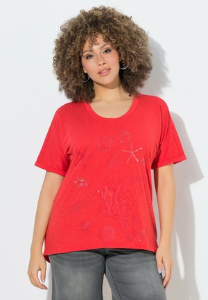 Frau mit lockigem Haar, die Creolen, ein rotes T-Shirt mit Meeresthema und graue Jeans trägt, steht vor einem weißen Hintergrund.