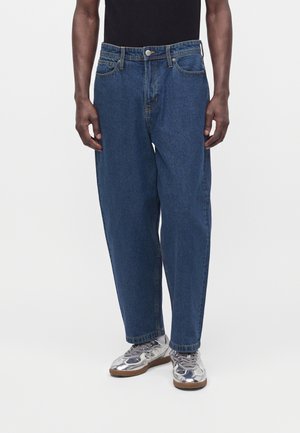Homme portant un jean bleu ample taille haute et des chaussures de sport argentées sur fond blanc.