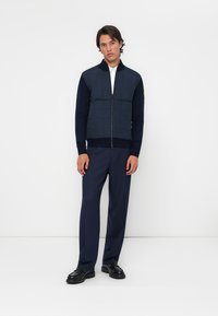 Veste bomber matelassée marine avec fermeture éclair à l'avant, poignets et col côtelés ; assortie avec un pantalon marine et des chaussures noires.