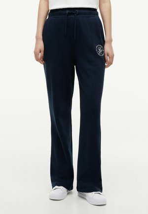 Persona che indossa pantaloni della tuta blu navy con coulisse, logo bianco sulla coscia sinistra e sneakers bianche, in piedi davanti a uno sfondo semplice.