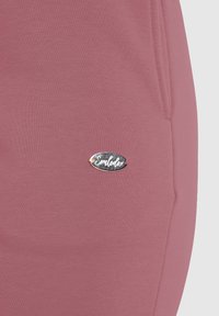 Rosa Sweatpants mit glatter Textur, ausgestattet mit einem silbernen ovalen Logo-Tag, das an der Seitenasche "Smilodox" zeigt.