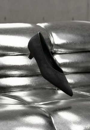 Black pointed-toe kitten heel shoe floating above a shiny metallic tufted leather couch.