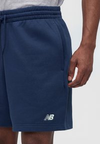 Tengerészkék sport rövidnadrág rugalmas derékpánttal, oldalzsebbel és fehér New Balance logóval a szegély felett egy személy combján.