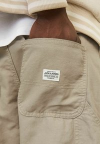 Pantalones cortos de cargo beige con un bolsillo frontal que tiene una etiqueta blanca visible que dice "JACK & JONES". Tela suave y duradera con detalles de costura visibles.