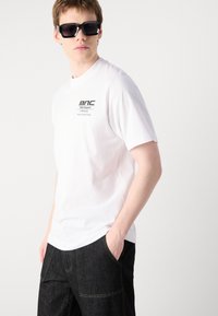 Hvit T-skjorte med kort erm som har en logo trykket på venstre bryst, kombinert med mørk denimbukser. Brukt med store svarte solbriller.