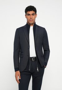 Blazer navy con collettino zip, due bottoni e tasche a pattina, abbinato a una camicia bianca e pantaloni scuri, creando un aspetto elegante.