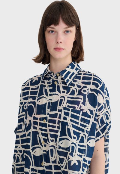 Jeune personne aux cheveux courts bruns portant une chemise boutonnée à manches courtes avec un motif bleu marine et blanc et une poche avant.