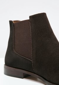 Botte Chelsea en daim brun foncé avec panneau latéral élastique et petit talon en bois sur fond blanc.