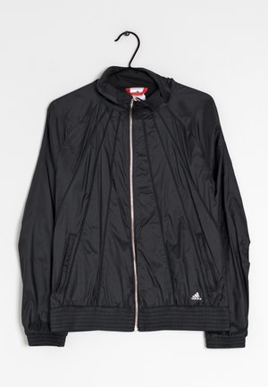 Veste noire Adidas à fermeture éclair, col montant, détails de coutures subtils et logo sur le bas du devant, suspendue à un cintre noir sur fond de mur blanc.