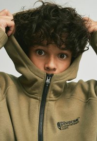 Khaki zielona bluza z zamkiem błyskawicznym i wysokim kołnierzem, czarnym zamkiem i logo, wykonana z teksturowanej, miękkiej tkaniny i zakończona ściągaczami.