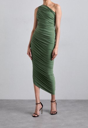 Kvinde iført en rynket, olivengrøn midi-kjole med ét skulderstykke, kombineret med sorte ankelstrop-sandaler med høje hæle, stående på en grå gulv.