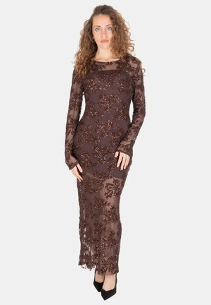 Donna con capelli ricci che indossa un abito lungo a maniche lunghe marrone trasparente decorato con paillettes floreali e tacchi neri a punta.