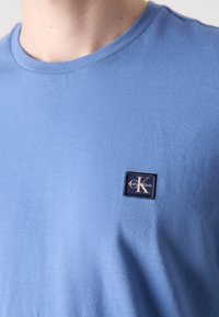 Világoskék pamut póló kerek nyakkivágással, kisebb, sötétkék téglalap alakú logópatccsal, amelyen a "Calvin Klein" felirat és egy stilizált "K" található.