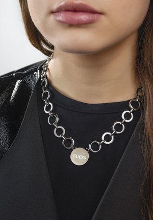 Gros plan sur une personne portant un collier chaîne argentée avec de grosses mailles et un pendentif rond gravé du mot "GUESS" sur un haut noir.