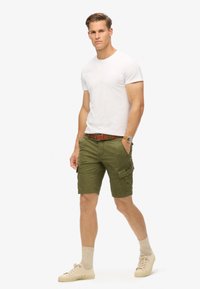 Pantaloni cargo verdi con molte tasche, una cintura marrone e una maglietta bianca. Sneaker crema e calzini chiari completano l'outfit.